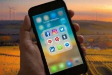 Hand hält Smartphone mit Social-Media-Apps, im Hintergrund eine dörfliche Landschaft