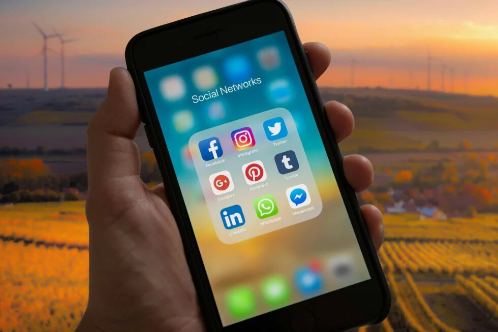 Social Media im ländlichen Raum, Symbolbild Hand hält Smartphone mit Social-Media-Apps, im Hintergrund eine dörfliche Landschaft